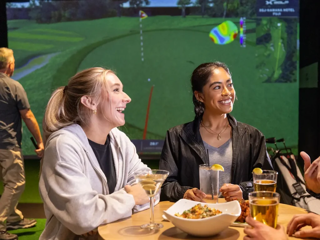 Two women smiling and chatting at a table with drinks and food during a bachelorette party at X-Golf, with a golf simulator and course details in the background.