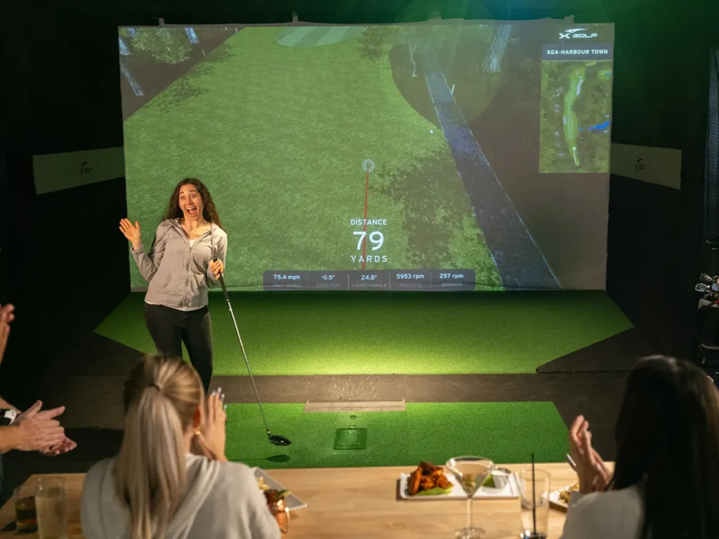 A woman celebrating a successful shot on an indoor golf simulator during a bachelorette party at X-Golf, with friends applauding from a table in the foreground.