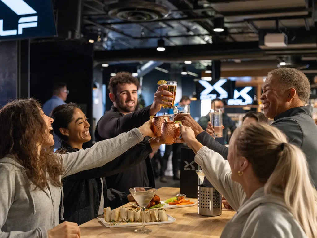 A group of friends raising their glasses for a toast at a birthday party hosted at X-Golf, with food and drinks on the table and the 'X Golf' logo in the background.