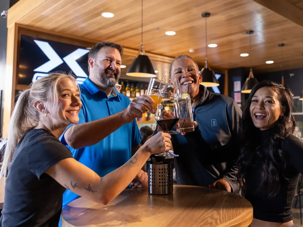 Smiling colleagues raising their glasses for a toast during a corporate event at X-Golf, standing near a table with the 'X Golf' logo visible in the background.