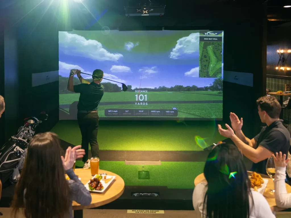 An attendee swinging a golf club on an indoor simulator during a corporate event at X-Golf, with colleagues applauding and enjoying food and drinks at nearby tables.