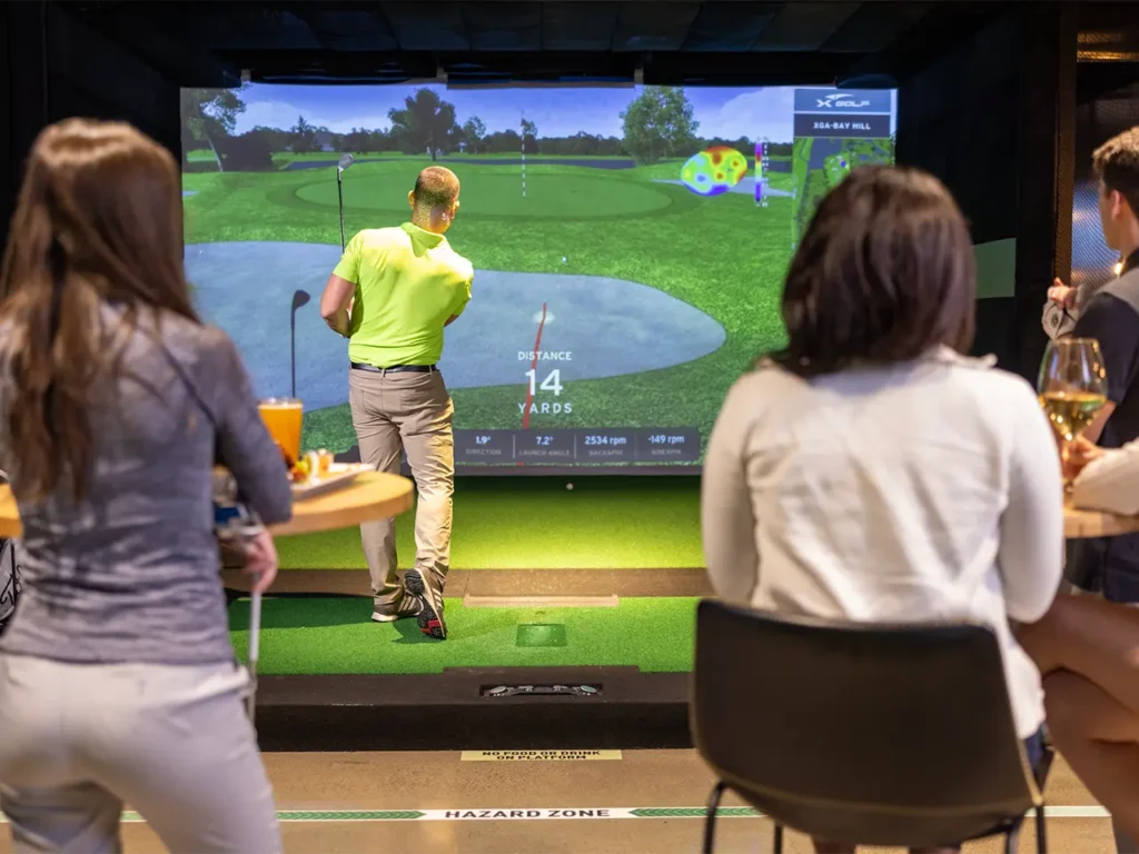 Man taking a golf shot on an indoor simulator during a fundraising event hosted at X-Golf, as attendees watch from nearby tables with food and drinks.