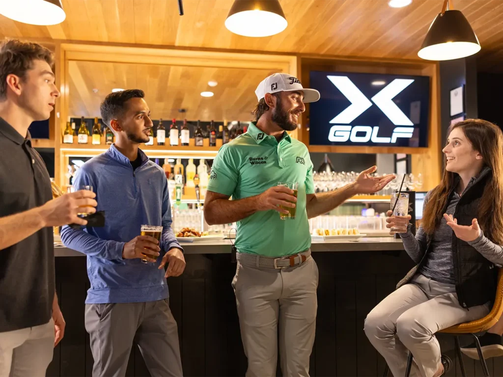 Attendees enjoying drinks and conversation at a fundraising event hosted at X-Golf, with the bar and 'X Golf' logo visible in the background.