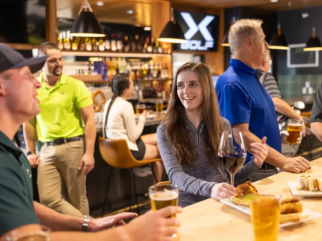 A lively bar scene at an 'X Golf' facility, with people enjoying drinks and food, including a woman smiling while holding a glass of wine.