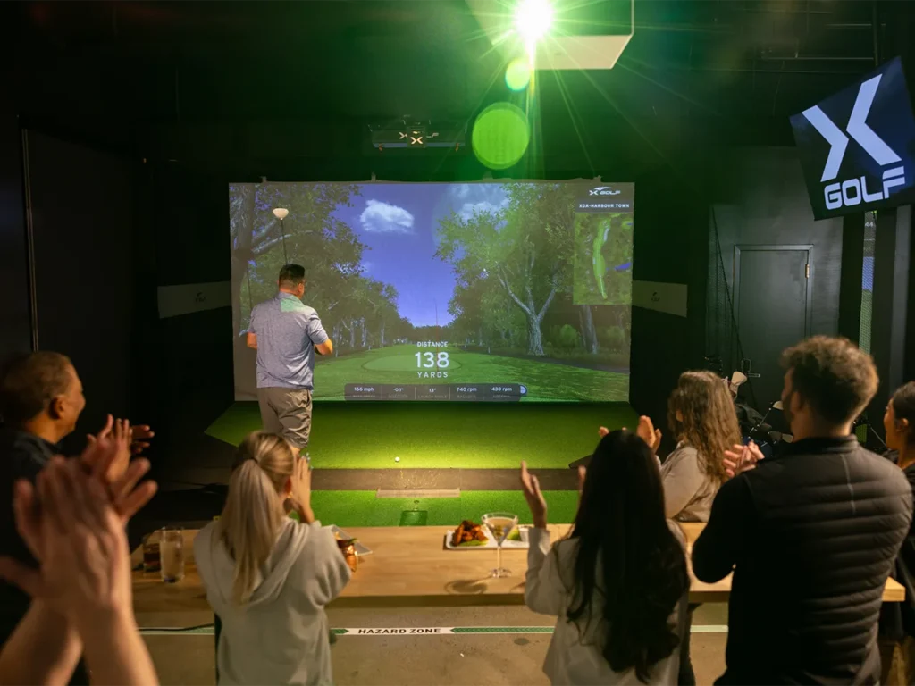 Group of people applauding as a man takes a shot in an indoor golf simulator, with 'X Golf' branding displayed on the screen.