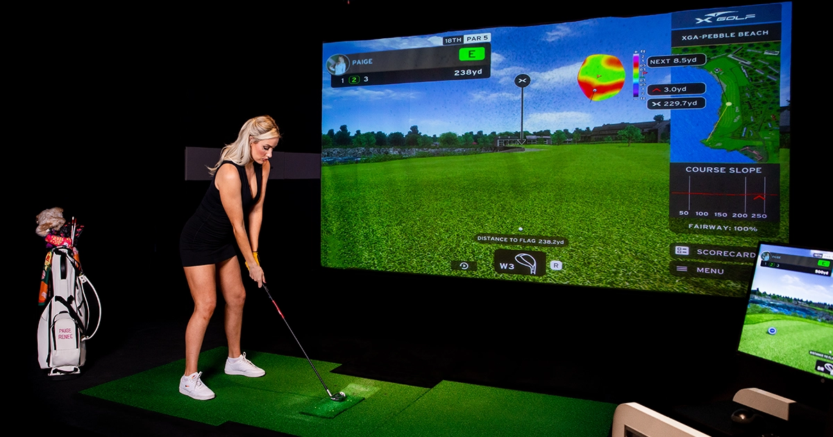 Paige Spiranac at X-Golf preparing to hit a golf ball on a simulator. The screen shows a virtual golf course with detailed metrics like distance to the flag and hole information.