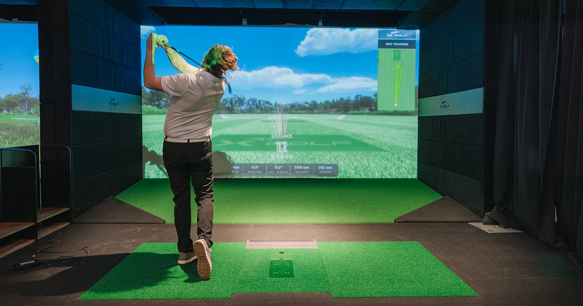 A person swings a golf club in an indoor simulator at X-Golf, with a virtual golf course displayed on the screen showing distance and trajectory data.