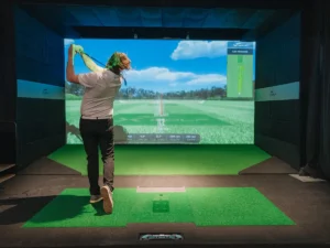 A person swings a golf club in an indoor simulator at X-Golf, with a virtual golf course displayed on the screen showing distance and trajectory data.