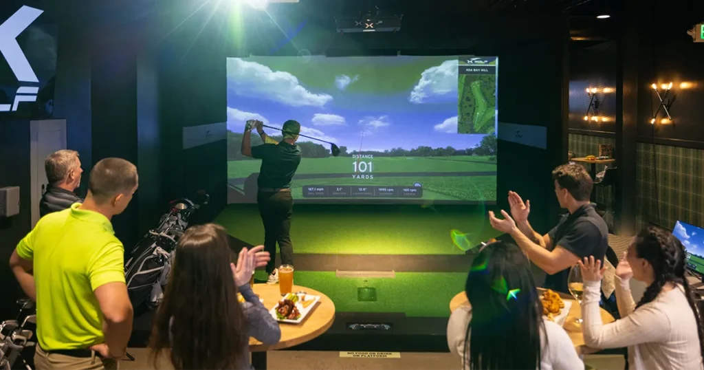 A person swings a golf club in front of an X-Golf screen while others clap and enjoy food at a corporate event.