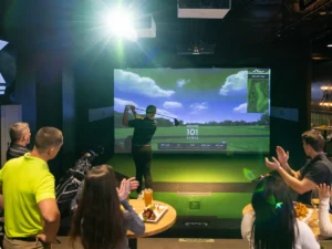 A person swings a golf club in front of an X-Golf screen while others clap and enjoy food at a corporate event.