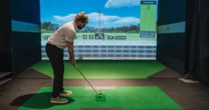 A man lines up a shot at an X-Golf indoor simulator, with virtual course and shot metrics displayed on a screen.