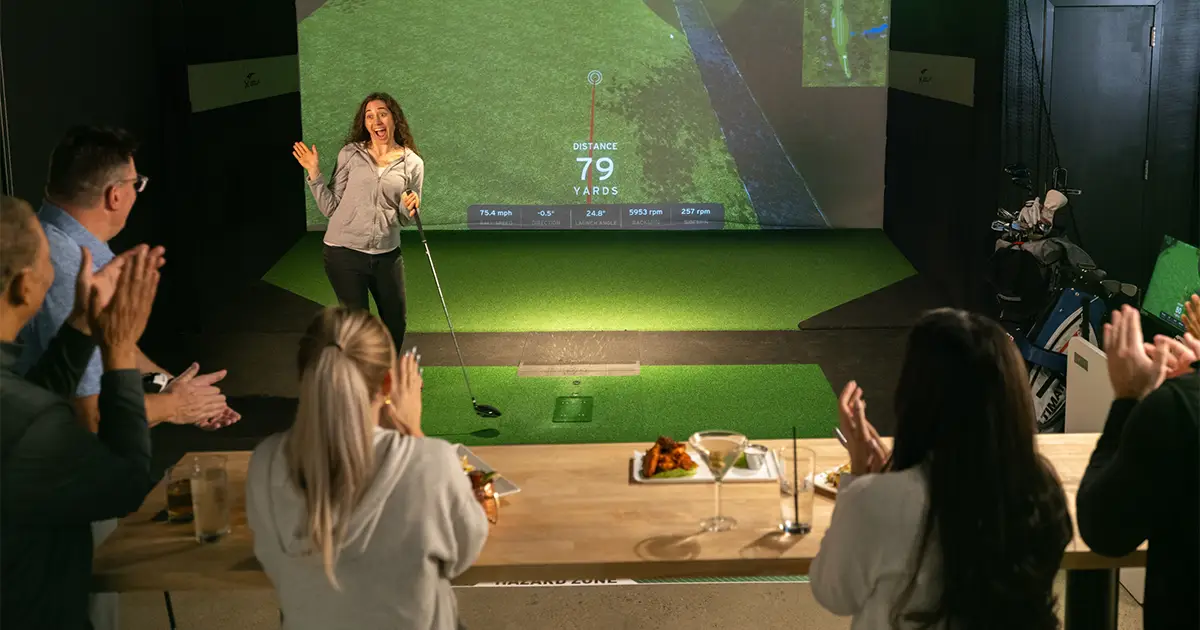 A woman celebrates a golf shot at an X-Golf simulator while a seated group applauds during a charity tournament event.