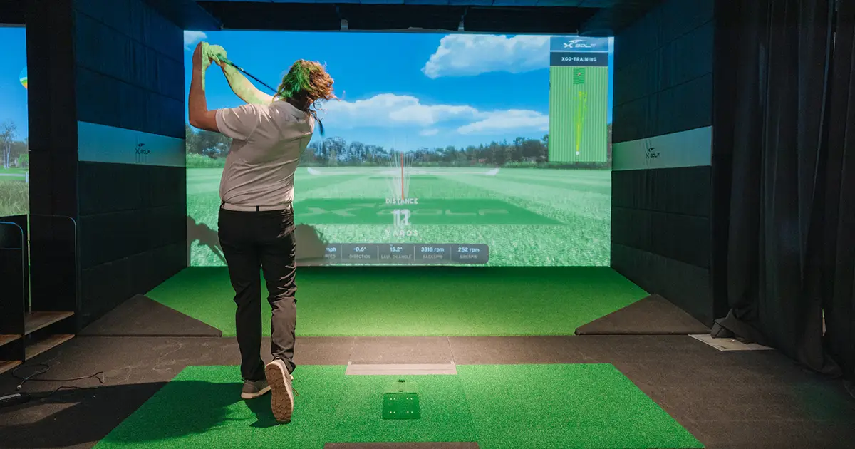 A man follows through on a swing at an X-Golf simulator while practicing basic drills for new golfers.