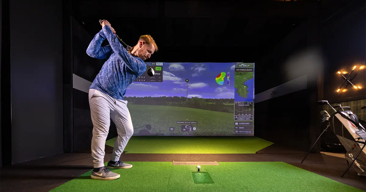 A man mid-backswing at an X-Golf simulator, with a virtual course and game stats projected on the screen.