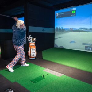 Man swinging golf club at a indoor simulator at X-Golf Frisco indoor simulator event space in Frisco texas. He is wearing funky pants and has white hair, showing that even unconventional to PGA players can be powerful.