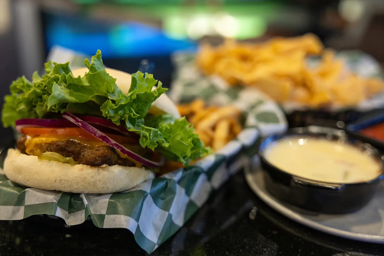 Juicy cheeseburger topped with lettuce, onions, and pickles, with fries and queso dip in the background.