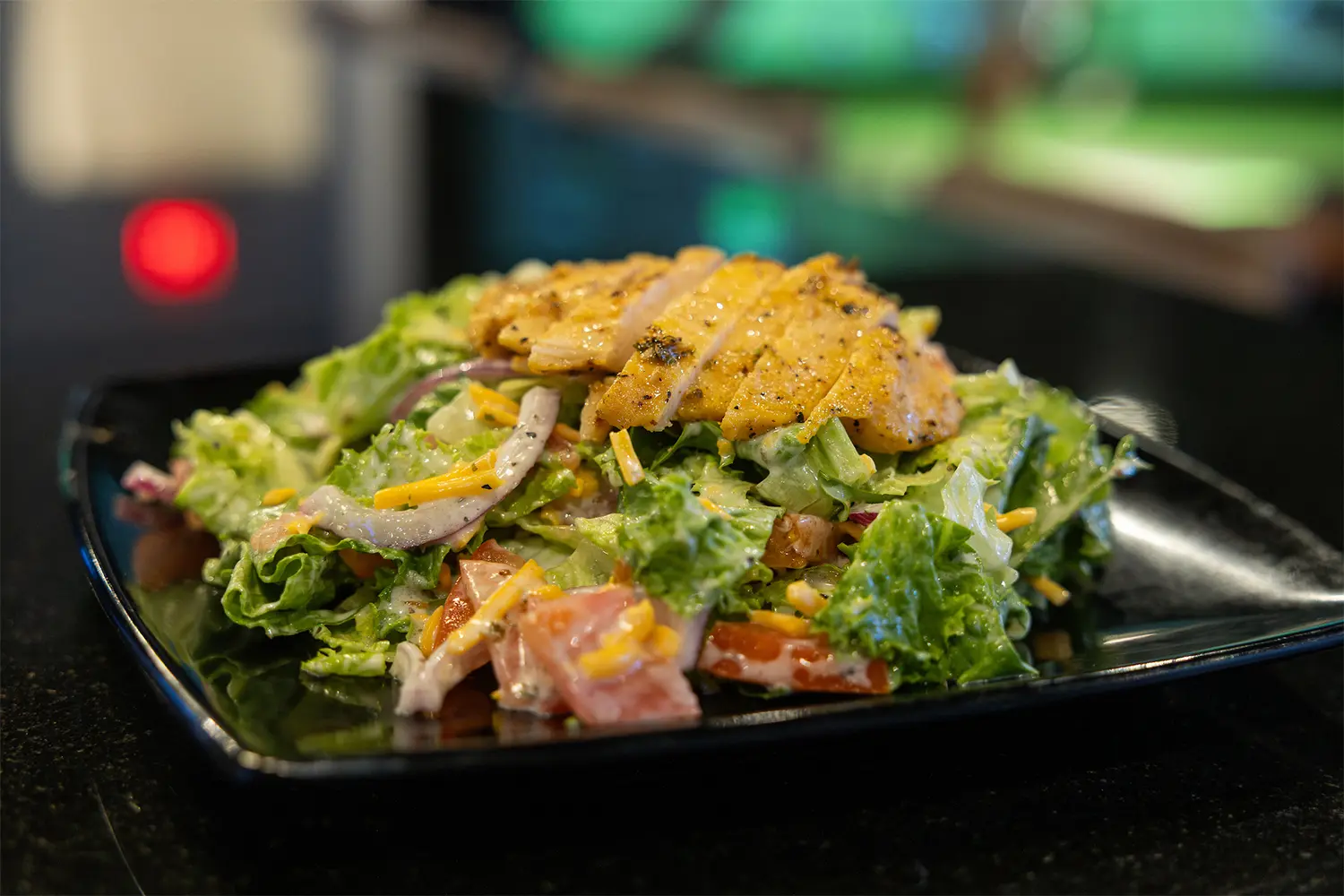 Fresh salad topped with grilled chicken, shredded cheese, onions, and tomatoes in a black square bowl.