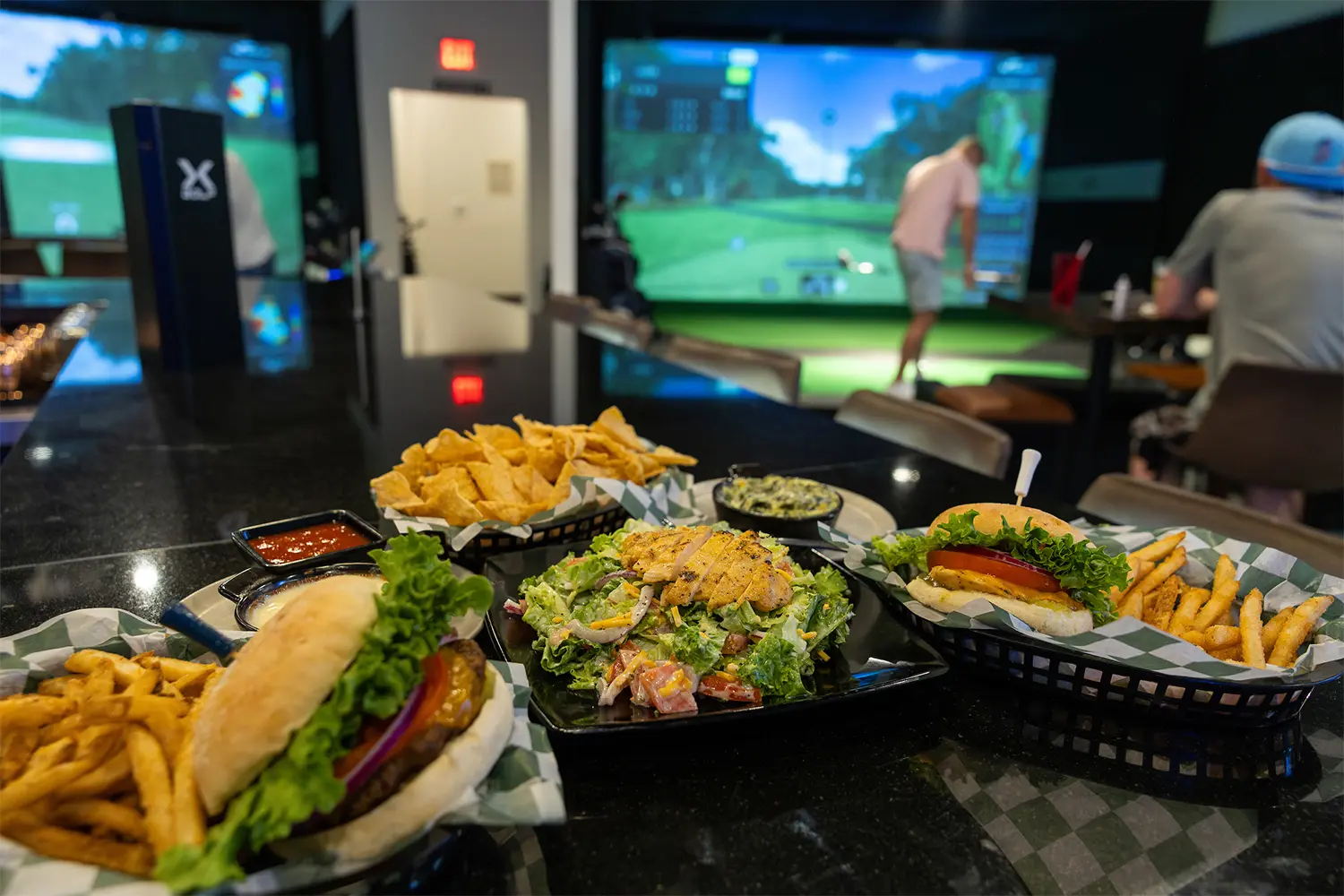 Plates of burgers, fries, chips, and salad on a bar counter with guests playing virtual golf in the background.