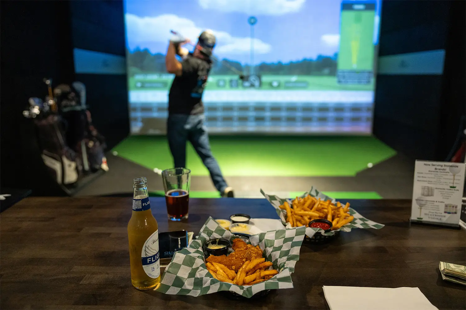 Basket of fries and chicken tenders on a table with a beer bottle in front of a golf simulator where a person is swinging a club.