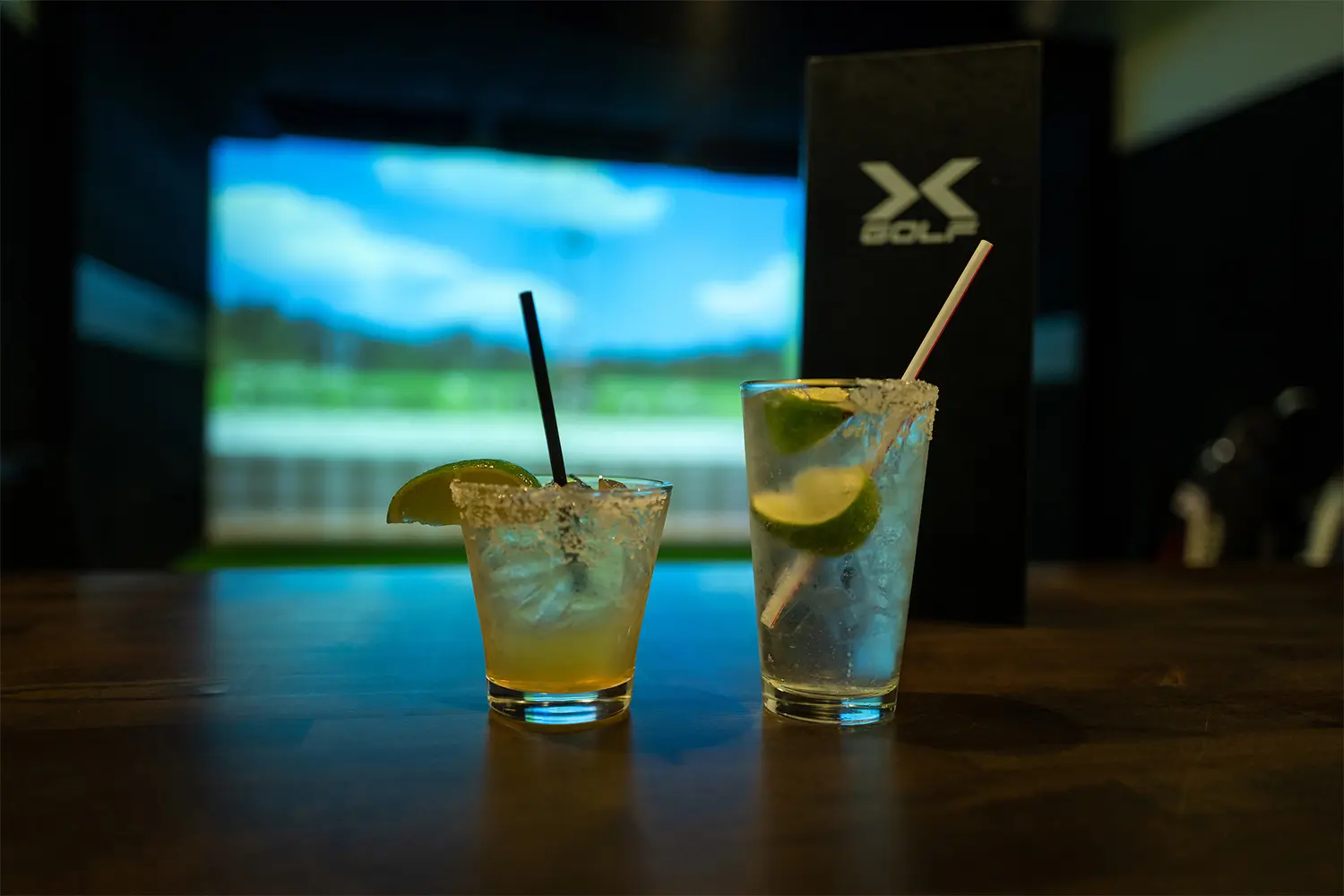 Two cocktails with lime wedges, one with a salted rim, set on a table with a golf simulator screen in the background.