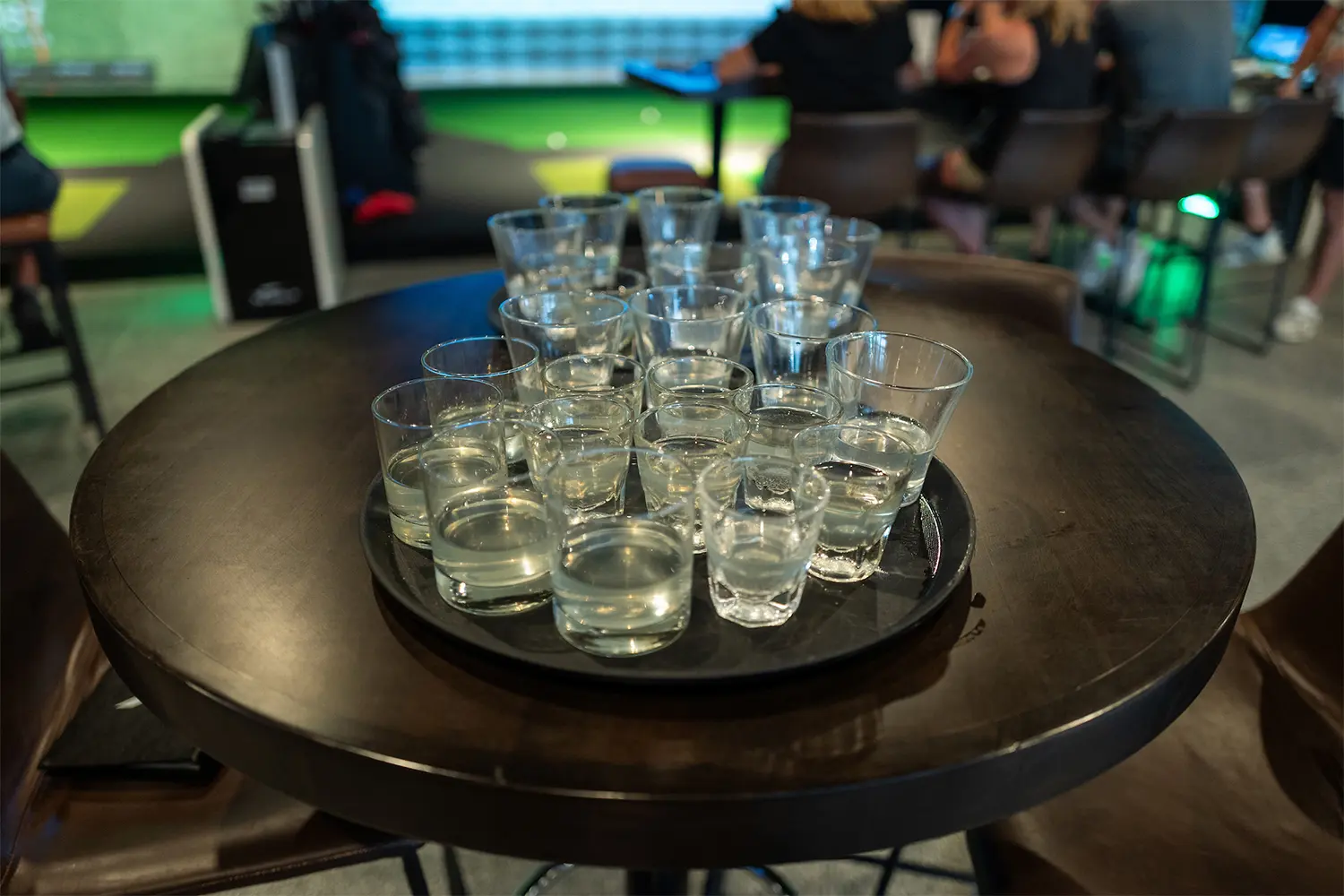 Tray of shot glasses filled with clear liquor on a round table at a golf simulator venue.