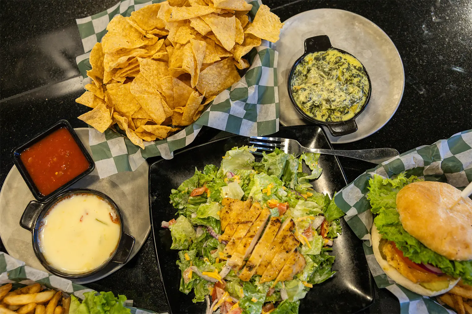 Top view of tortilla chips, dips, a chicken salad, and a sandwich arranged on a black countertop.