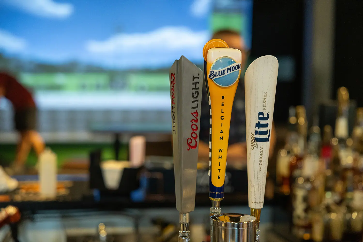 Beer taps for Coors Light, Blue Moon, and Miller Lite with a blurred golf simulator in the background.