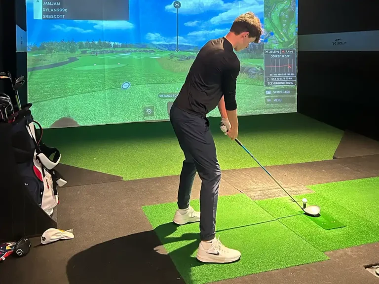 Golfer in black shirt addresses ball in X-Golf simulator bay. Screen shows team names JAMJAM, DYLAN9990, and GR8SCOTT.