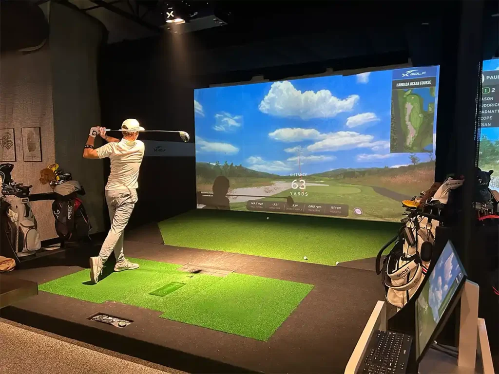 Golfer completes swing in X-Golf simulator bay. Screen displays 63-yard shot on Hanbada Ocean Course with ball flight data.