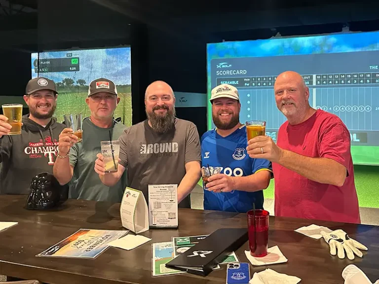 Five men raise drinks in a toast at an indoor golf simulator bar at X Golf Frisco, with an X Golf scorecard displayed on the screen behind them