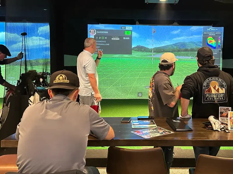 Three men watch a putt tracked on an indoor simulator screen displaying the Hanbada Ocean Course at X Golf Frisco, with scorecards and golf gear on the bar counter in the foreground